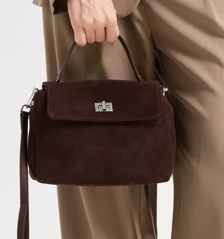 A dark brown suede handbag with a silver-tone interlocking 'G' clasp, held by a model wearing beige trousers against a light gray background.