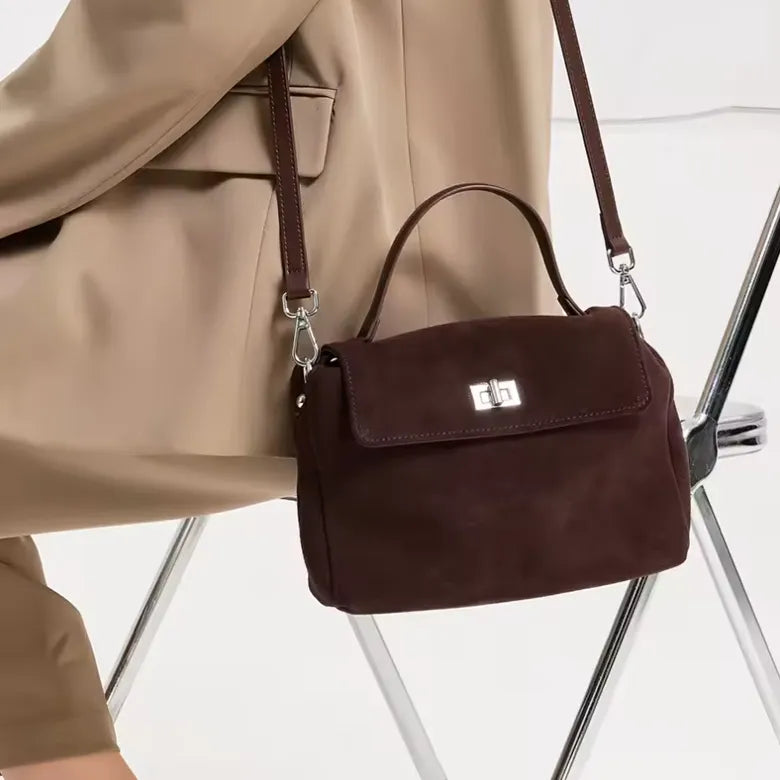 A dark brown suede handbag with a top handle and shoulder strap, featuring a silver turn-lock clasp on the front flap. The bag is shown worn crossbody over a beige blazer, with the legs of a white chair visible in the background.